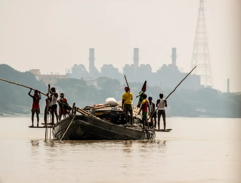 Fishermen poling a traditional boat on a river with an industrial skyline behind