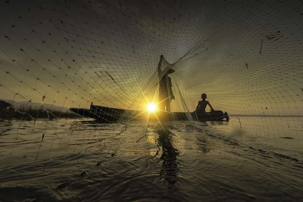 Silhouette of a fisherman casting a net from a boat at sunrise.