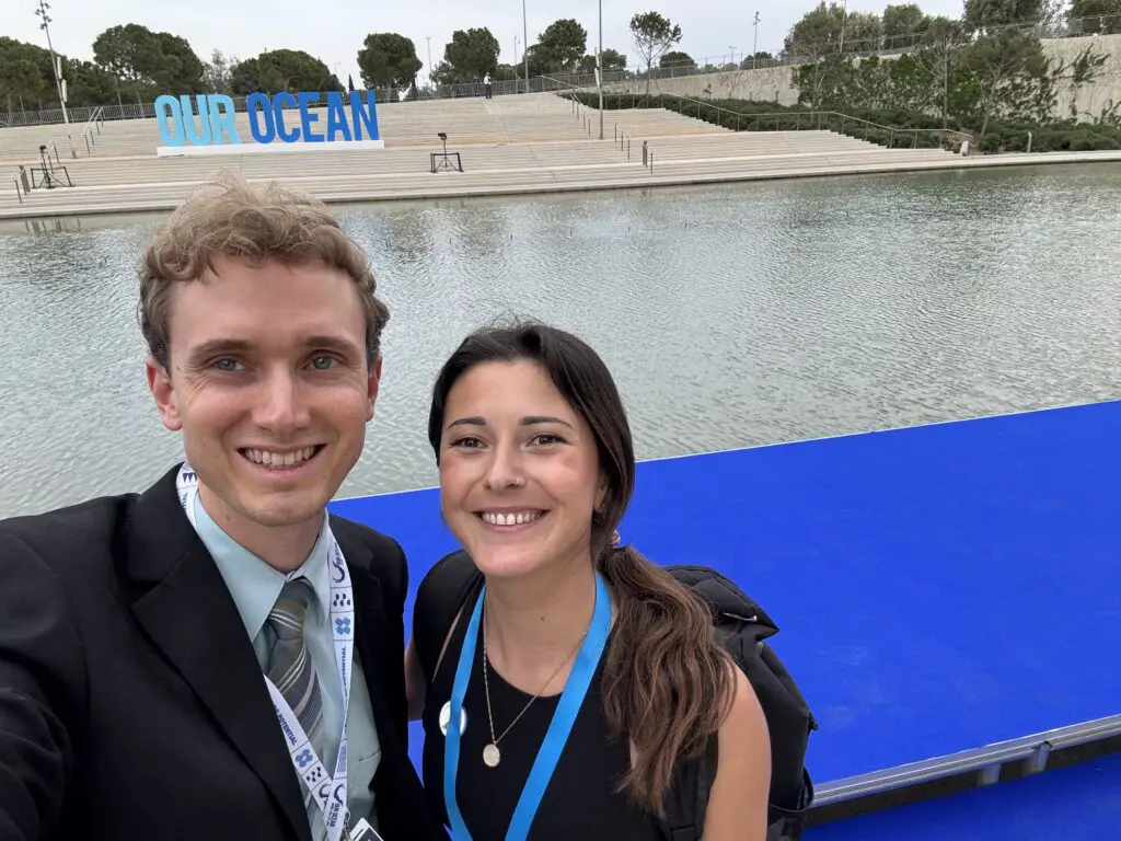 Man and woman taking a selfie in front of an Our Ocean sign by the water