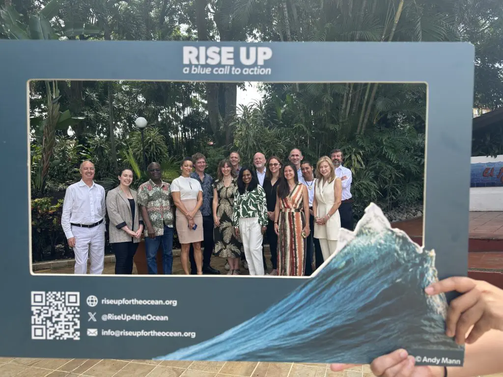 Group of about twelve people posing behind a Rise Up branded photo frame in a tropical garden