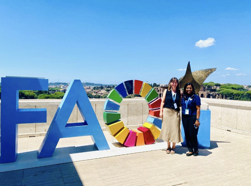Two women posing next to the FAO sign and SDG wheel sculpture at FAO headquarters in Rome