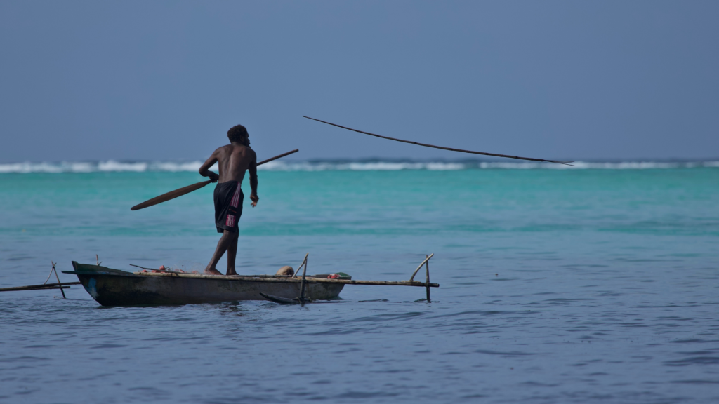 Fisher standing on an outrigger canoe in turquoise waters