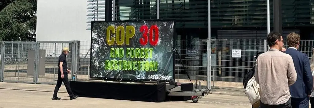 A close-up of a Greenpeace COP30 End Forest Destruction billboard on a trailer, with people walking past.