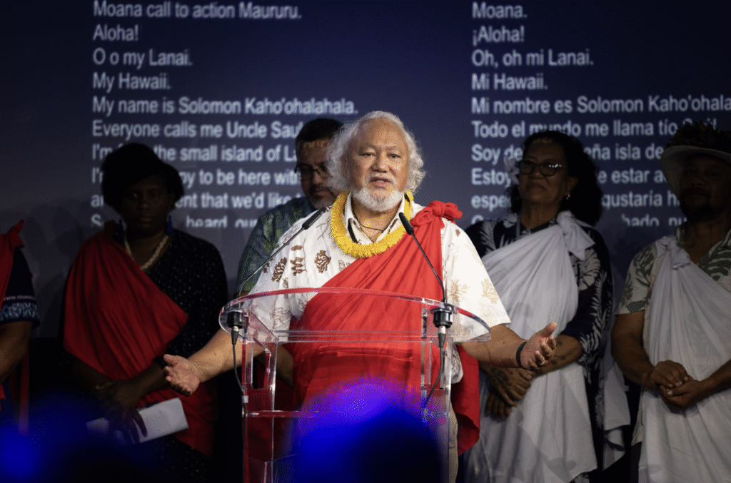 An older man in traditional dress speaking at a microphone in front of a screen showing song lyrics.