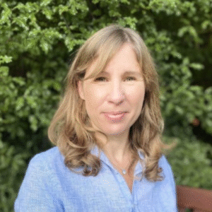 Headshot of a woman with wavy light-brown hair wearing a blue shirt, greenery behind her.