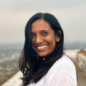 Headshot of a smiling woman with long dark hair in a white top, a city view behind her.