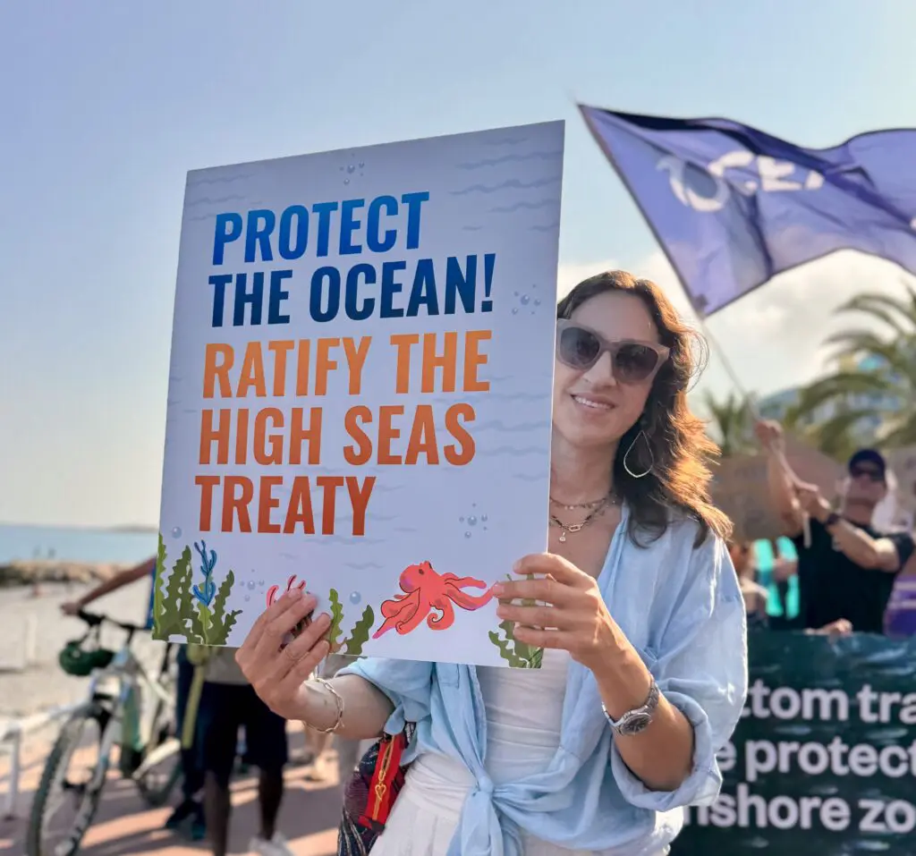 Woman holding a “Protect the Ocean, Ratify the High Seas Treaty” placard at a seafront march.