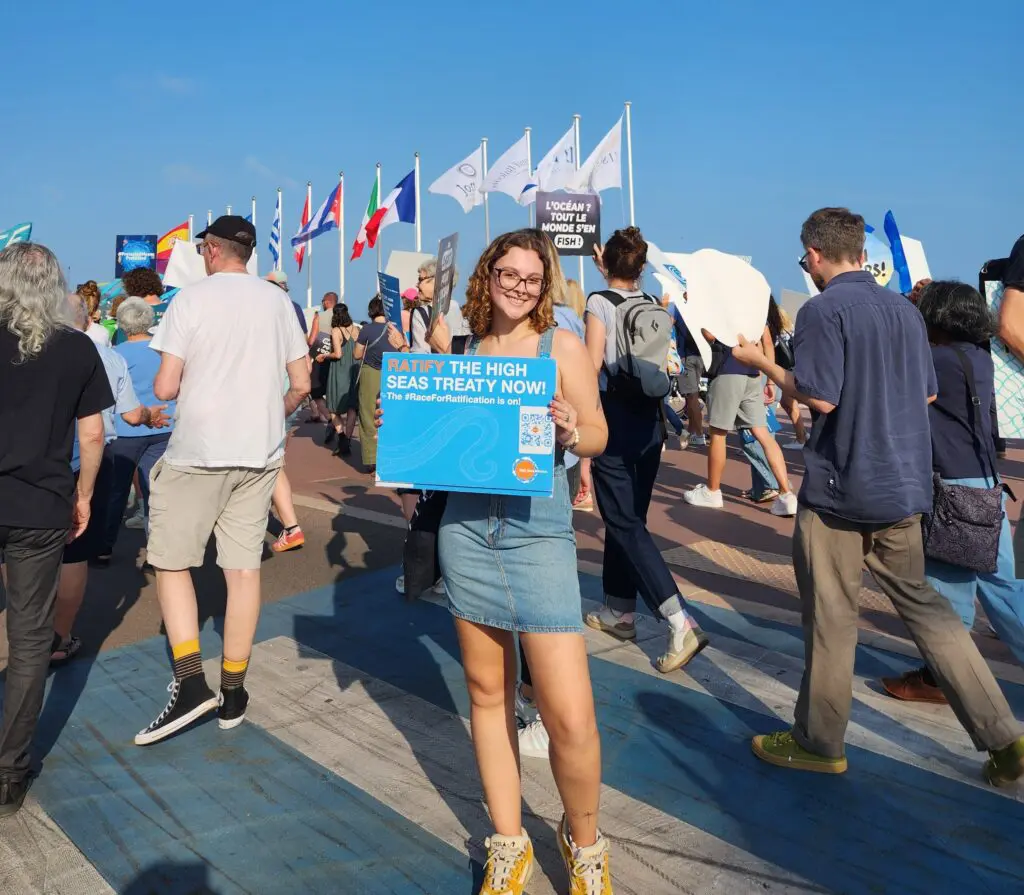 A woman holding a "High Seas Treaty Now" placard at an outdoor rally with flags