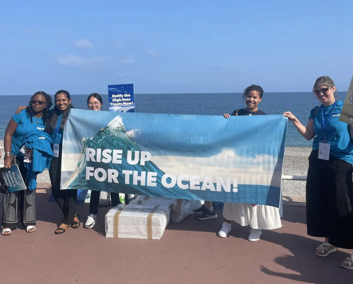Group in RISE UP T-shirts holding a large “RISE UP for the Ocean” banner by the sea.