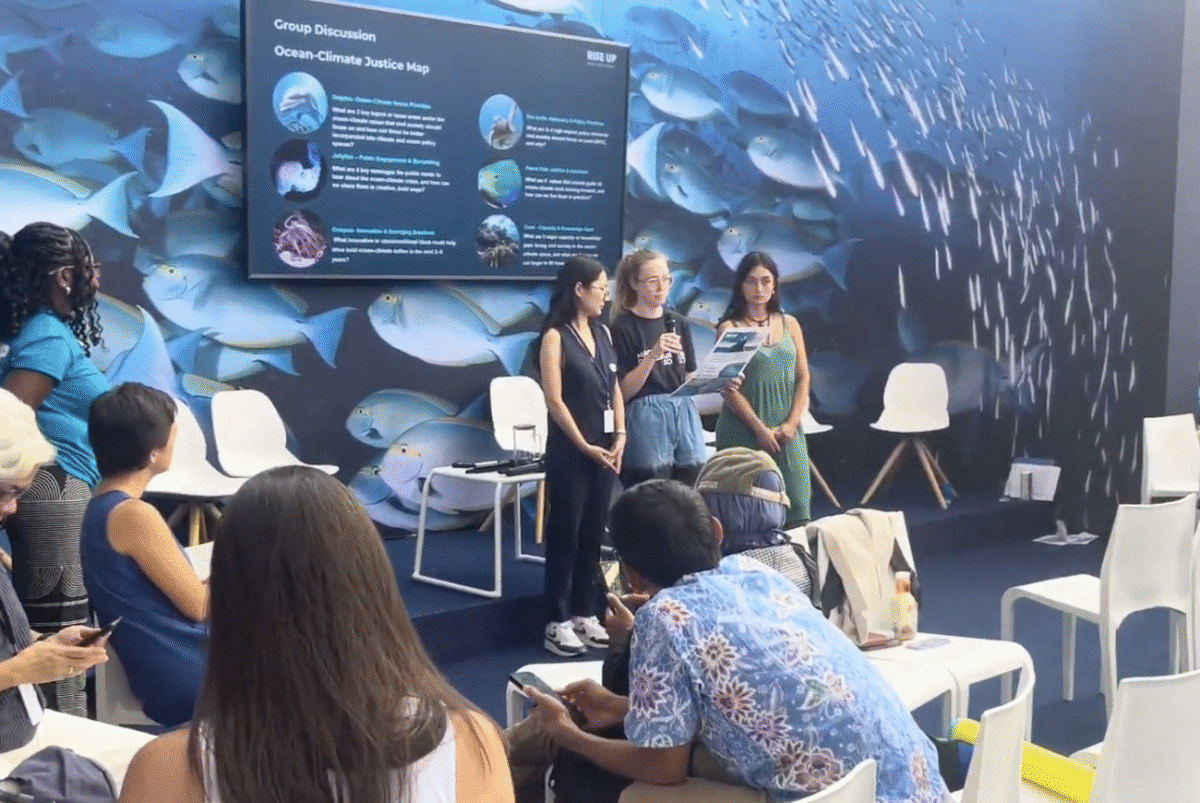 Presenters on stage speaking to a seated audience in front of a fish mural.