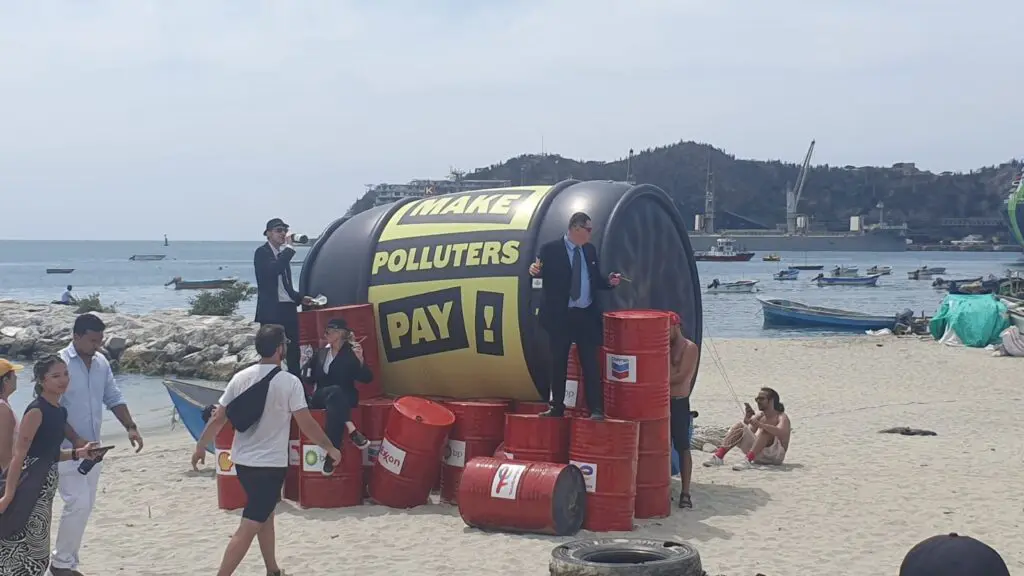 Activists in business suits stand on a giant inflatable oil drum reading “Make Polluters Pay”, surrounded by red oil barrels branded with fossil fuel company logos, on the beach at Santa Marta.