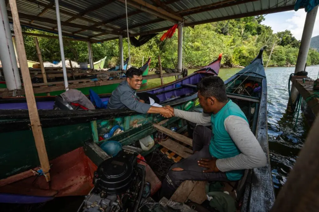 Two men shaking hands across the side of traditional fishing boats moored beside mangroves