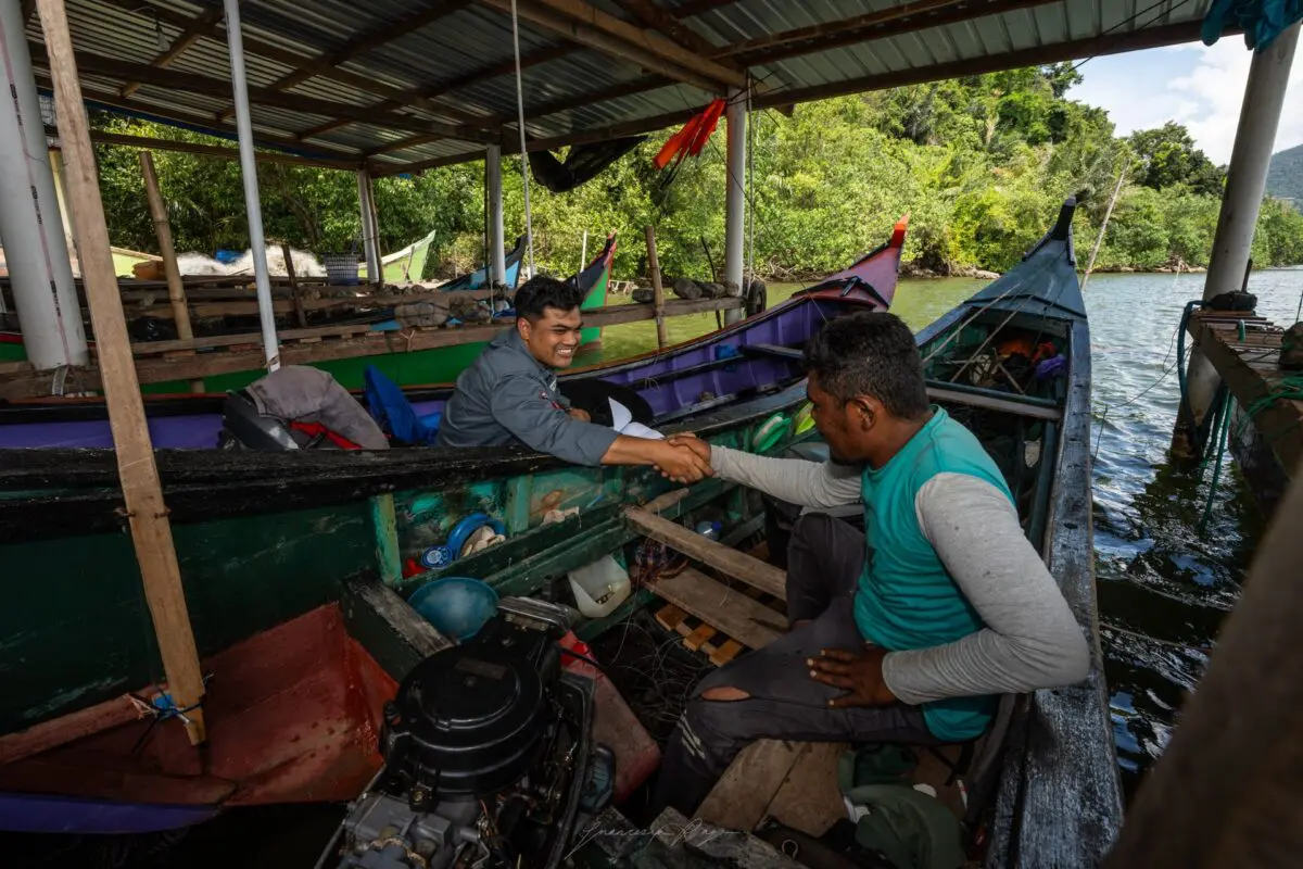 Two men shaking hands across the side of traditional fishing boats moored beside mangroves