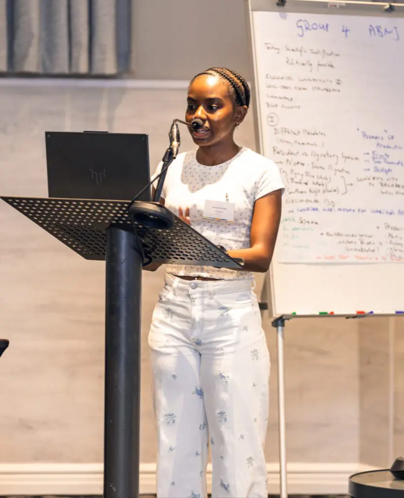 Woman speaking at a lectern beside a whiteboard covered in workshop notes