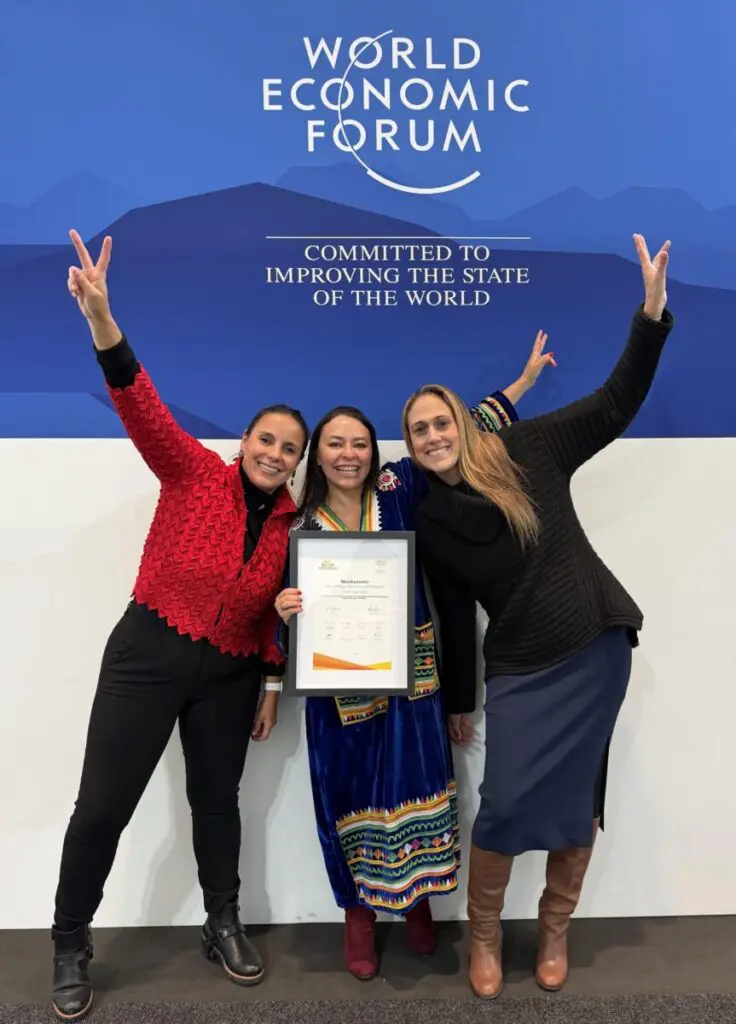 Three women cheering with raised hands while holding a framed award in front of a World Economic Forum backdrop
