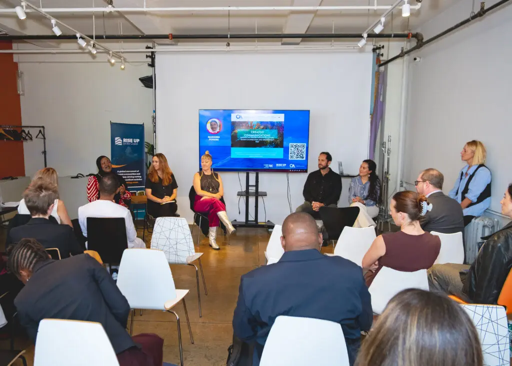 Wide view of the storytelling panel: five panellists seated facing an audience, beside a RISE UP banner and a screen showing a 'Creative Communications' slide