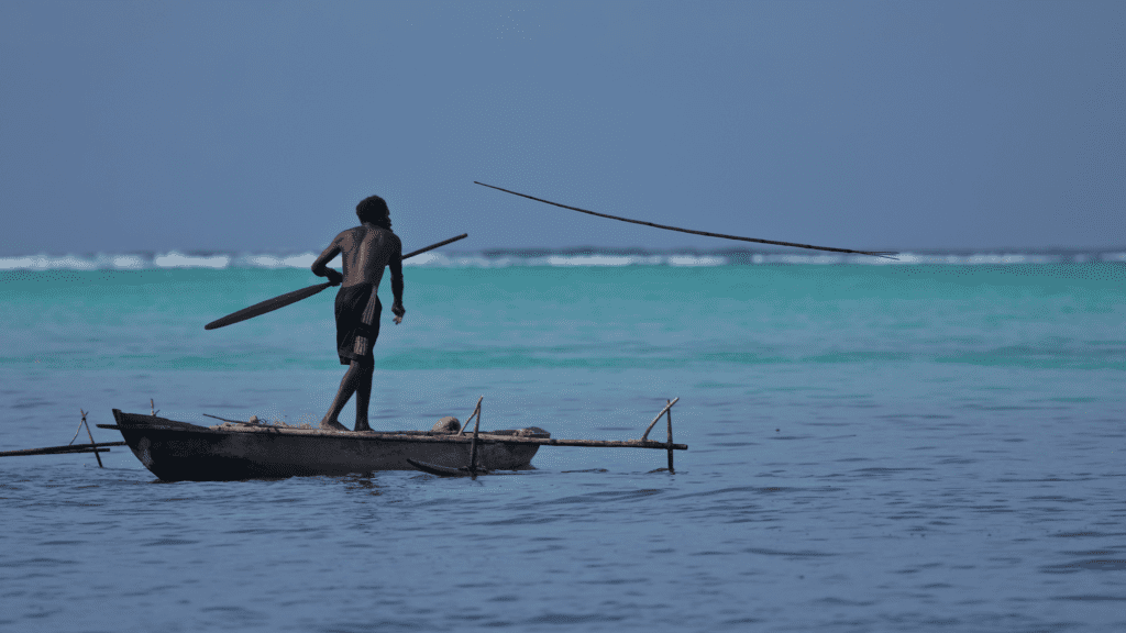 Fisher standing on an outrigger canoe in turquoise waters