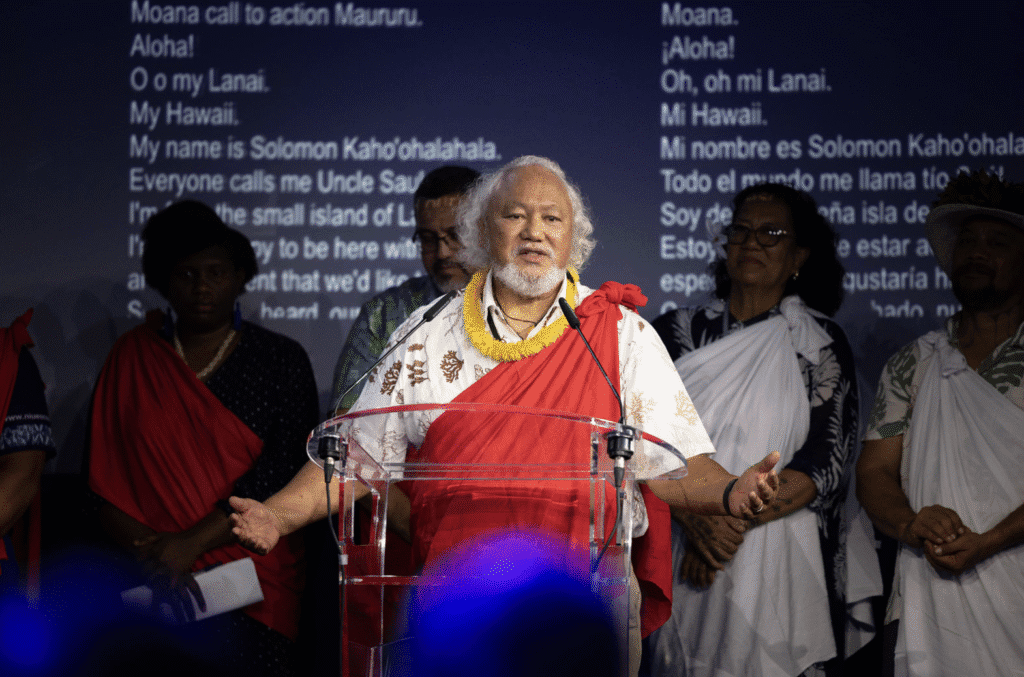 An older man in traditional dress speaking at a microphone in front of a screen showing song lyrics.