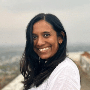 Headshot of a smiling woman with long dark hair in a white top, a city view behind her.