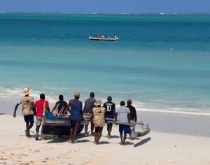 A group of people carrying a wooden outrigger fishing canoe up a white sand beach, with another small boat out on the turquoise water behind them.