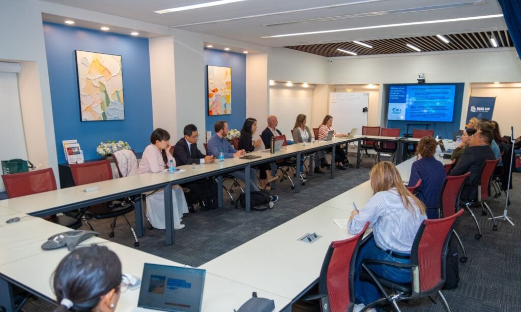 Participants seated around a U-shaped conference table at the Ocean, Energy and Communities session, with a CIEL presentation on screen and a RISE UP banner